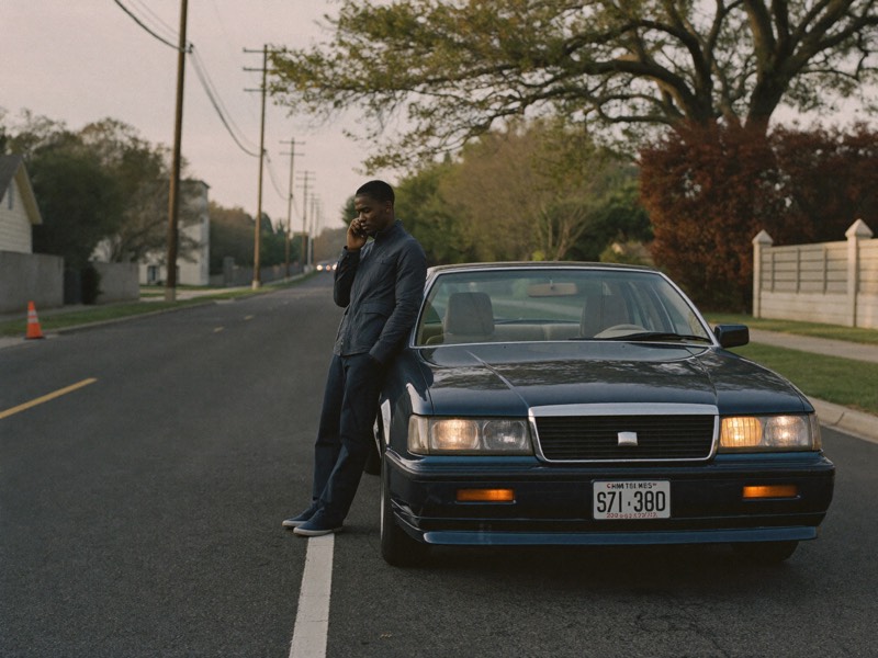 A driver stands beside their car on a quiet road, making a phone call.